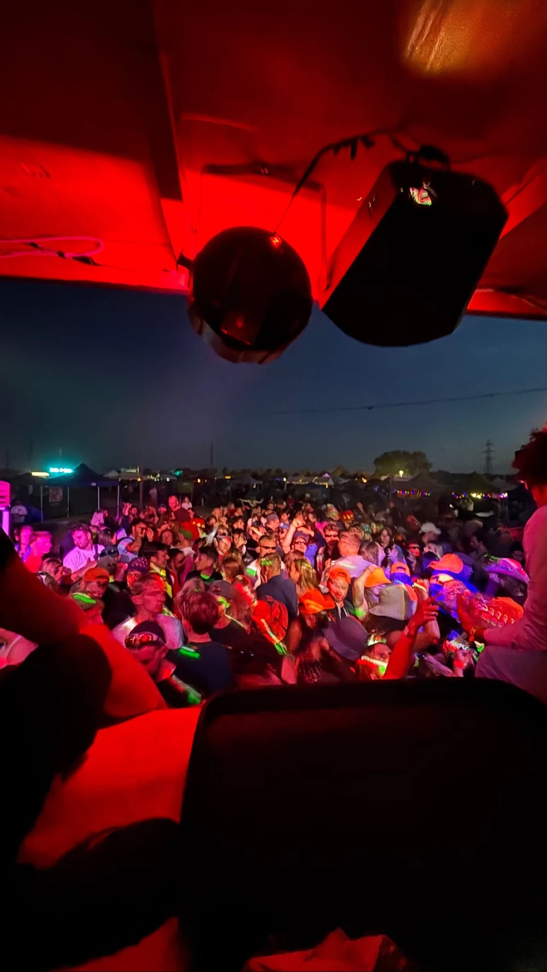 Festivalpublikum under en rød overdækning med højttaler og diskokugle, set fra DJ-pulten.